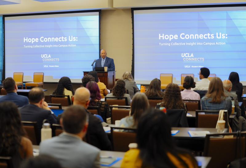 A man speaking in front of a large group of people inside a conference room.