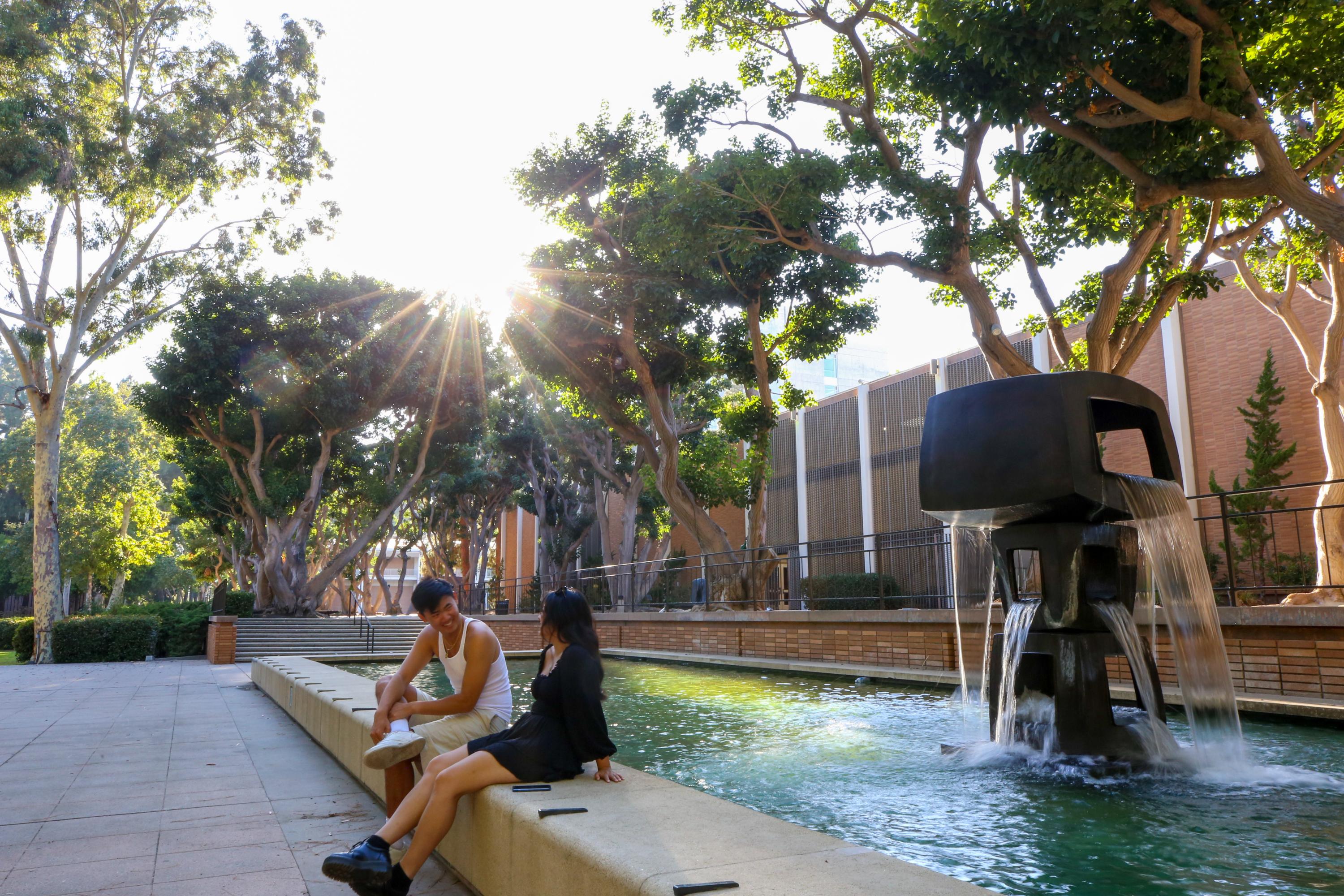 Two students sitting next to each other at a fountain on a sunny day at the UCLA campus.