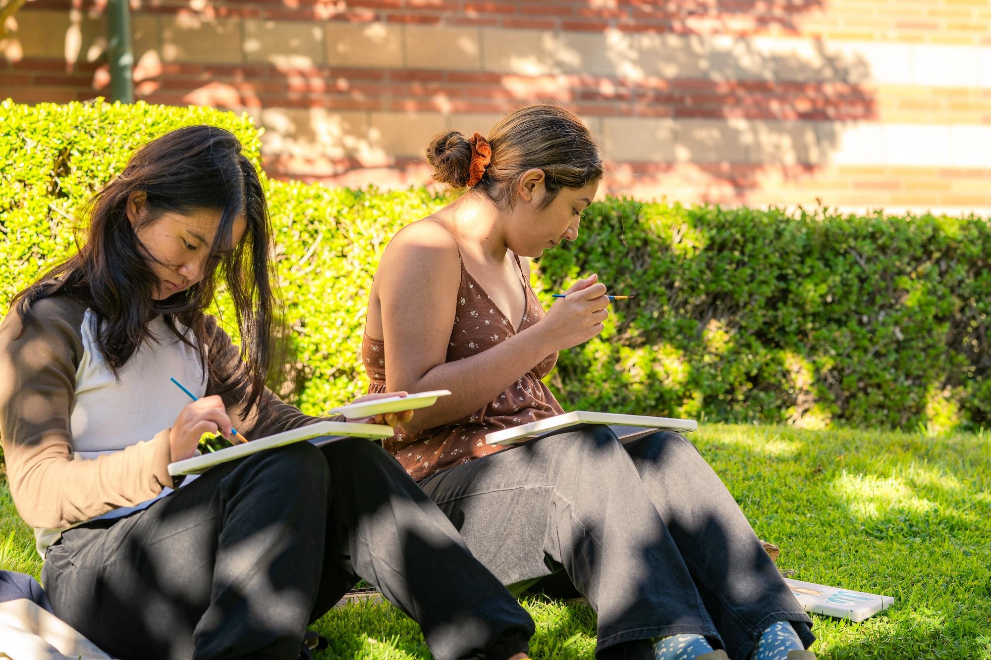 Two students painting while sitting on the grass on a sunny day.