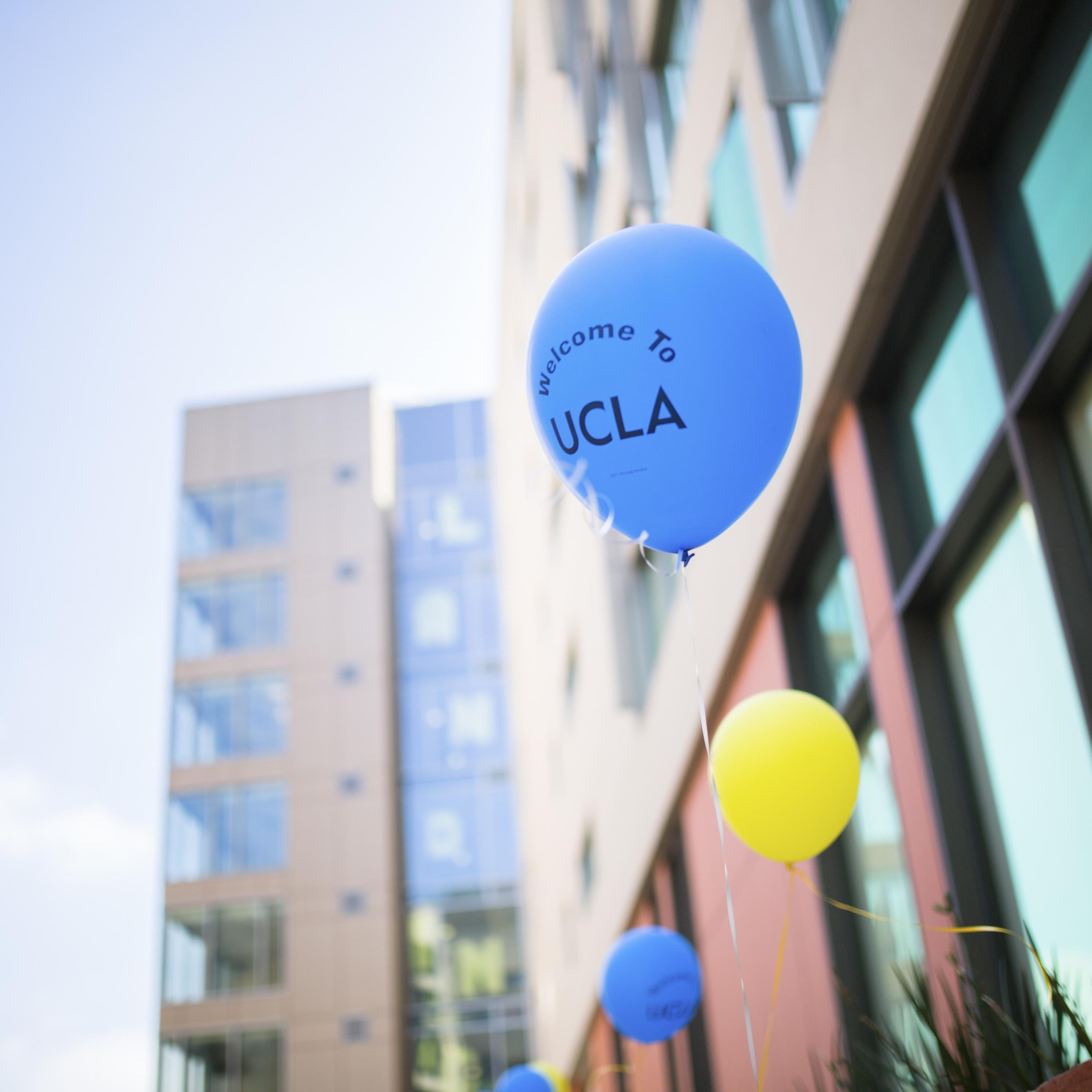 Blue and yellow balloons that say "Welcome to UCLA" float in front of modern buildings