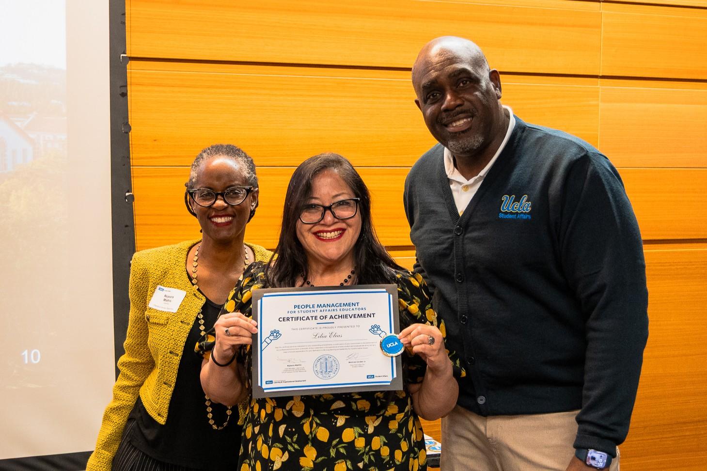 Two women and one man smiling at the camera. Woman in the middle is holding a certificate of completion for a workshop series.