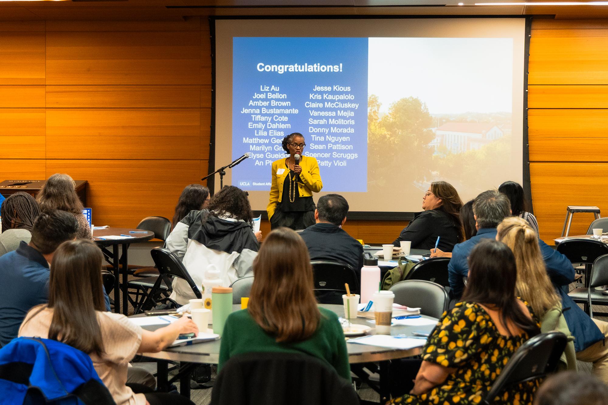 Photo of a woman speaking into a microphone in front of a group of people.