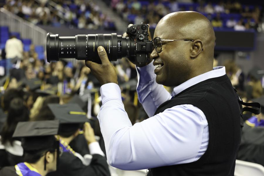 A man holding a large camera in a crowd, taking photos of a UCLA graduation ceremony.